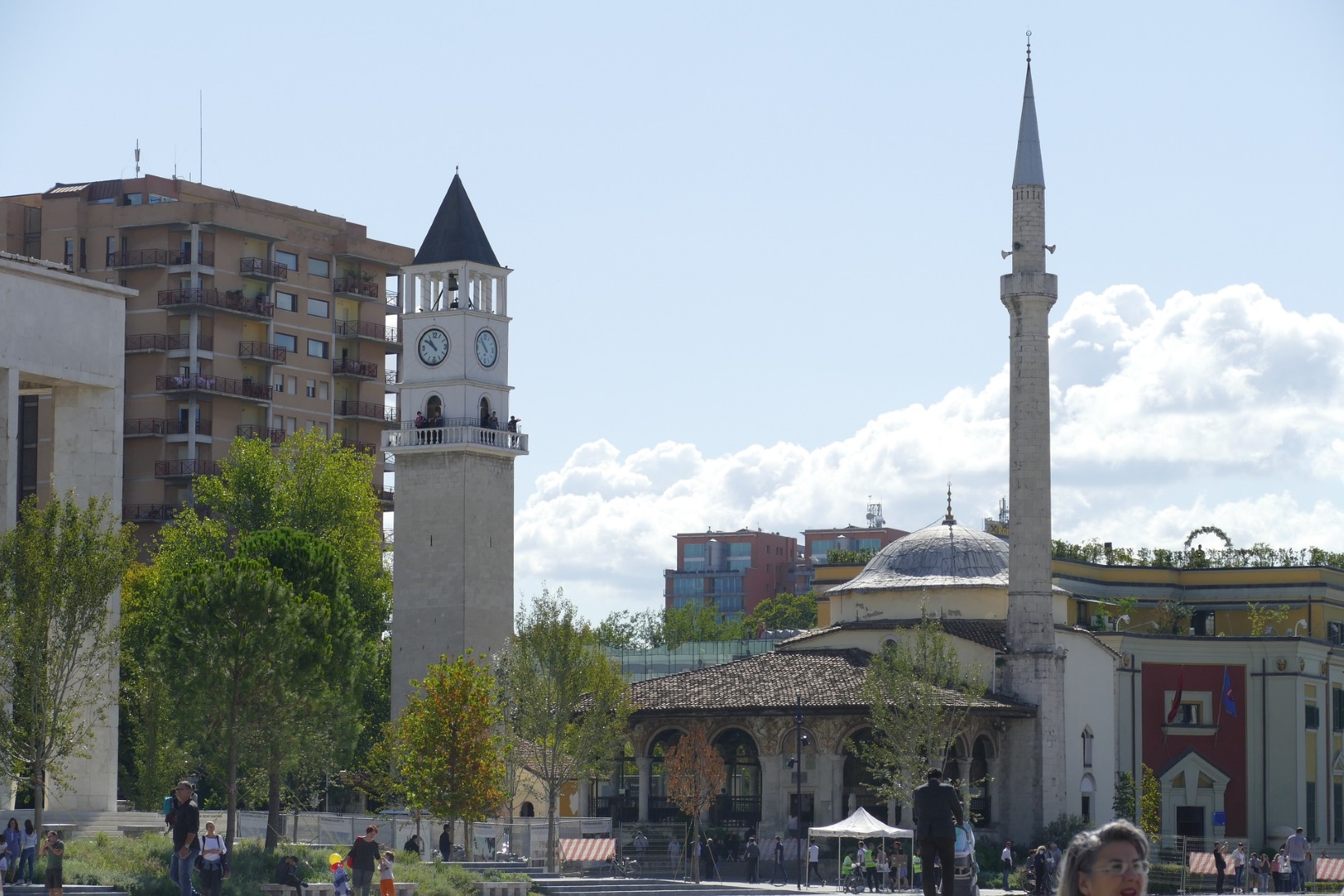 La Torre del Reloj y la mezquita Et'hem Bey de Tirana (Albania) se erigen como importantes hitos históricos en medio de edificios modernos y una exuberante vegetación bajo un cielo resplandeciente.